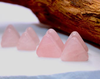May include: Several pink rose quartz pyramids are arranged on a white surface. The pyramid shapes are smooth and translucent, with a soft, rosy hue. A piece of brown wood is in the background.