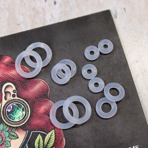 May include: A collection of translucent, circular silicone O-rings in various sizes. These flexible, clear rings are arranged in a cluster, showcasing their smooth, round profile and hollow centers, designed for use as jewelry components or seals.