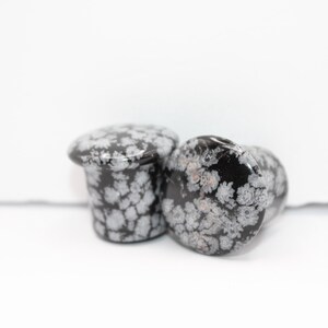 These Snowflake Obsidian Double Flared Plugs feature a smooth, polished surface with natural gray-white crystal "snowflakes" scattered across a rich black volcanic glass base. The double flare ensures a secure, comfortable fit for stretched ears with