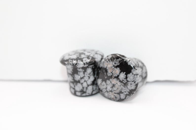 These Snowflake Obsidian Double Flared Plugs feature a smooth, polished surface with natural gray-white crystal "snowflakes" scattered across a rich black volcanic glass base. The double flare ensures a secure, comfortable fit for stretched ears with