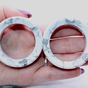 May include: Two white marble-patterned bangle bracelets. The bracelets are circular with a smooth, polished finish. The marble design features gray veining against a white background. The bracelets are held in a hand.
