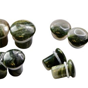 A striking pair of Green Jasper Single Flare Plugs is displayed against an earthy-toned backdrop. Each plug features deep forest green hues with natural patterns of dark veining and subtle swirls, unique to every piece. The surface is smoothly polish