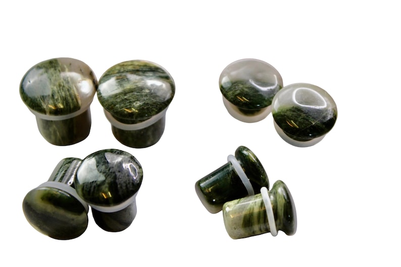 A striking pair of Green Jasper Single Flare Plugs is displayed against an earthy-toned backdrop. Each plug features deep forest green hues with natural patterns of dark veining and subtle swirls, unique to every piece. The surface is smoothly polish
