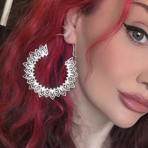 May include: Silver hoop earrings with a detailed, openwork floral design. The earrings are large and circular, with a delicate, symmetrical pattern. The earrings are worn against a backdrop of vibrant red hair.