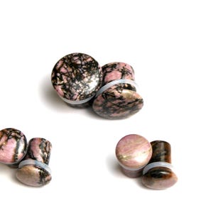 May include: A collection of stone ear plugs in various sizes. The plugs feature a pink and black marbled pattern. Each plug has a clear, rubber O-ring for secure placement. The plugs are displayed on a white background.