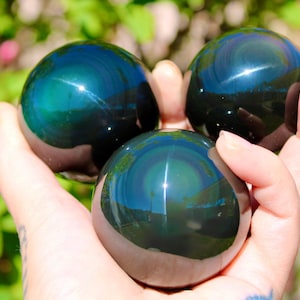 Könnte beinhalten: Drei irisierende Obsidiankugeln, jede mit einer polierten, reflektierenden Oberfläche. Die Kugeln zeigen eine Reihe von Farben, darunter tiefes Blau, Grün und Goldtöne. Die Kugeln werden in einer Hand gehalten, mit einem verschwommenen grünen Hintergrund.