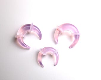 Puede incluir: Tres expansores de oreja translúcidos en forma de media luna, de diferentes tamaños. Los expansores tienen un degradado de rosa y morado, con un anillo transparente en el centro. Los objetos se muestran sobre un fondo blanco.