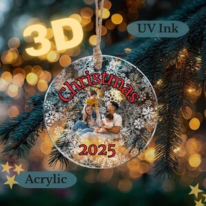 May include: A clear acrylic Christmas ornament with a family photo inside, surrounded by snowflakes. The word "Christmas" is in red script, and the year "2025" is below the photo. The ornament hangs from a twine loop.