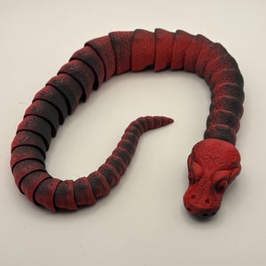 Puede incluir: Un juguete de serpiente articulada roja y negra. El juguete está hecho de piezas segmentadas que le permiten doblarse y moverse. La cabeza de la serpiente está detallada con ojos y una superficie texturizada.