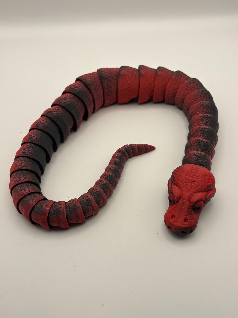 May include: A red and black articulated snake toy. The toy is made of segmented pieces that allow it to bend and move. The head of the snake is detailed with eyes and a textured surface.
