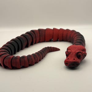 May include: A red and black articulated snake toy. The toy is made of segmented pieces, allowing it to bend and move. The head is a solid red color, and the body transitions from black to red.