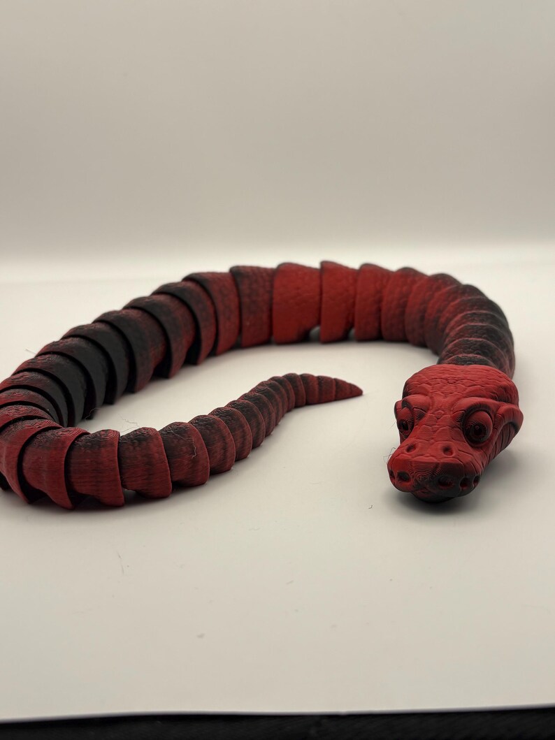 May include: A red and black articulated snake toy. The toy is made of segmented pieces, allowing it to bend and move. The head is a solid red color, and the body transitions from black to red.