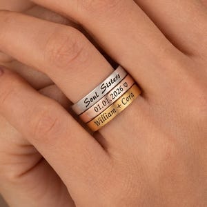 Op de afbeelding: Een stapel van drie dunne metalen ringen in zilver-, roségoud- en goudtinten. Elke ring is voorzien van zwarte gegraveerde tekst: de zilveren ring toont Soul Sisters, de roségouden ring 01.01.2026 met een hartje, en de gouden ring William + Cora.