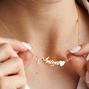 May include: A gold-colored necklace with the name "Anya" in cursive script, followed by a small heart-shaped charm. The necklace is being held up by a person wearing a white shirt.