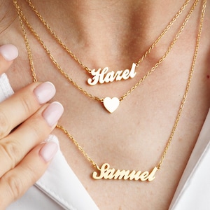 Puede incluir: Tres collares dorados con cadenas finas. El collar superior deletrea "Hazel", el del medio tiene un colgante de corazón y el inferior dice "Samuel". Los collares se usan sobre una camisa blanca.