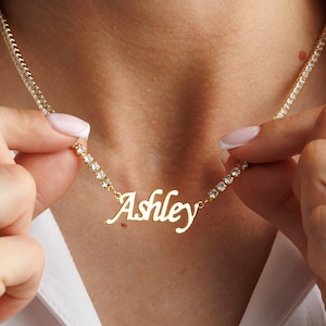 May include: A gold necklace spelling "Ashley" in cursive. The chain is adorned with small, clear gemstones. The person is wearing a white collared shirt. The necklace is being held up.