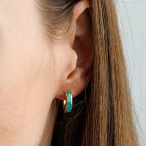 May include: Gold-colored hoop earrings with a turquoise inlay. The earrings are small and round, with a bright, polished finish. The turquoise inlay adds a pop of color and texture to the design. The earrings are shown on an ear.