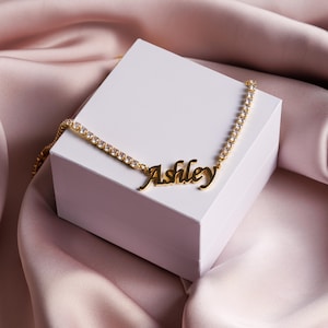Op de afbeelding: Een goudkleurige ketting met een tennis-kettingontwerp voorzien van heldere, sprankelende stenen en een centrale naamplaat-hanger met de naam Ashley in een cursief lettertype, rustend op een witte vierkante doos.