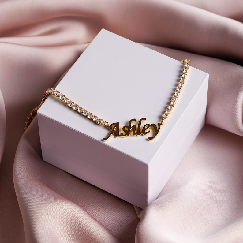 Op de afbeelding: Een goudkleurige ketting met een tennis-kettingontwerp voorzien van heldere, sprankelende stenen en een centrale naamplaat-hanger met de naam Ashley in een cursief lettertype, rustend op een witte vierkante doos.