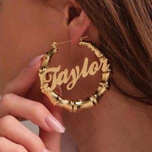 May include: A gold-toned hoop earring featuring a bamboo-inspired texture and a central script nameplate that reads Taylor. The circular earring has a polished metallic finish and a thin post closure, designed to be worn as a statement accessory.