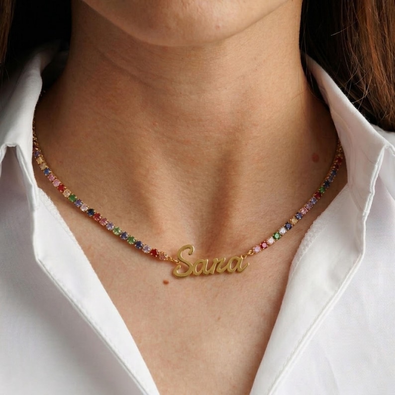 Op de afbeelding: Een goudkleurige ketting met een doorlopende streng van kleine, veelkleurige ronde edelstenen. In het midden van de ketting hangt een goudkleurige naamplaatje met de naam Sana in een vloeiend, cursief lettertype geschreven.