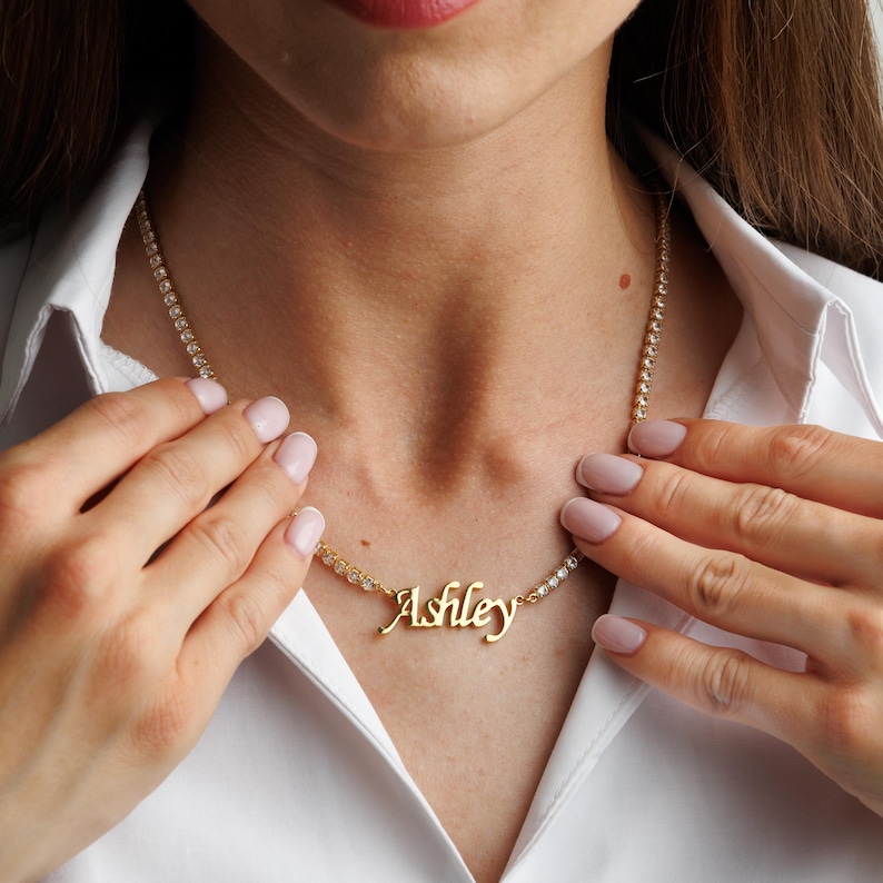 Op de afbeelding: Een goudkleurige naamketting met de naam Ashley in een cursief lettertype, gecentreerd aan een tennisarmbandketting bestaande uit heldere, rondgeslepen stenen. De ketting wordt gedragen over een wit overhemd met kraag, wat de afwerking benadrukt.