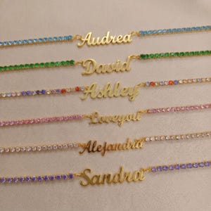 Op de afbeelding: Zes goudkleurige naamkettingen met hangers in cursief schrift. Elke ketting is versierd met kleine, vierkante kristallen in blauw, groen, veelkleurig, roze, transparant en paars, passend bij de namen Audrey, David, Ashley, Loveyou, Alejandra en Sandra.