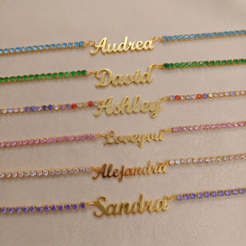 Op de afbeelding: Zes goudkleurige naamkettingen met hangers in cursief schrift. Elke ketting is versierd met kleine, vierkante kristallen in blauw, groen, veelkleurig, roze, transparant en paars, passend bij de namen Audrey, David, Ashley, Loveyou, Alejandra en Sandra.