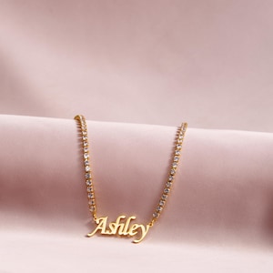 Op de afbeelding: Een goudkleurige ketting met een tennis-kettingontwerp en heldere, sprankelende stenen. In het midden van de ketting bevindt zich een goudkleurige naamhanger met de naam Ashley in een vloeiend, cursief lettertype.