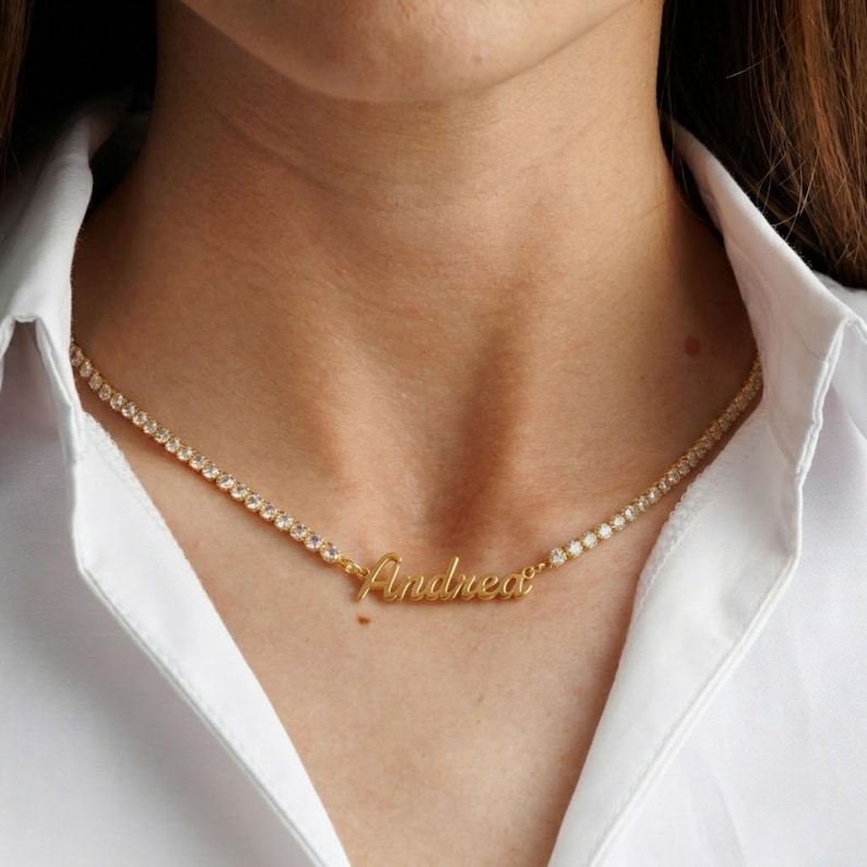 Op de afbeelding: Een goudkleurige tennis-halsketting met een doorlopende rij heldere, rondgeslepen stenen. In het midden van de ketting bevindt zich een goudkleurige naamhanger met de naam Andrea in een cursief lettertype, ge&iuml;ntegreerd in de ketting.