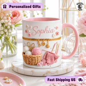 Puede incluir: Una taza de cerámica blanca con interior y asa de color rosa, que presenta el nombre Sophia en letra cursiva rosa. La taza está decorada con adornos en relieve que incluyen una cesta tejida con ovillos de lana, agujas y botones.