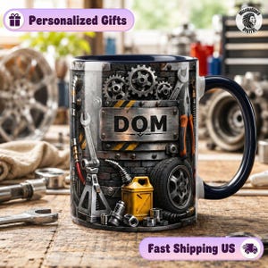 May include: A ceramic mug with a dark blue handle and interior, featuring a graphic print of a mechanical workshop scene with gears, wrenches, a car tyre, a yellow oil can, and a central metal plate displaying the name DOM in bold black letters.