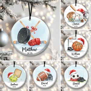 May include: Round Christmas ornaments with sports themes. One features hockey equipment with the name "Matthew" and the year "2025". Other ornaments show baseball, basketball, tennis, football, and soccer, each with a name and the year "2025".