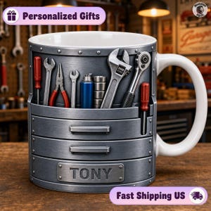 Pode incluir: Uma caneca de cerâmica projetada para se parecer com uma caixa de ferramentas de metal com interior e alça brancos. O exterior apresenta um acabamento cinza metálico com detalhes moldados de várias ferramentas manuais, incluindo chaves, alicates e chaves de fenda. Uma placa retangular na frente está gravada com o nome TONY.