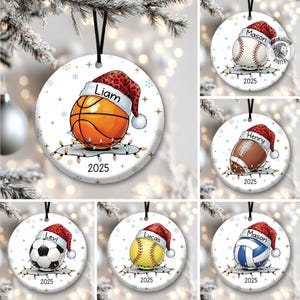 May include: A collection of round Christmas ornaments featuring various sports balls, each adorned with a Santa hat and the name of a person. The ornaments include a basketball, baseball, football, soccer ball, softball, and volleyball, all with the year 2025.
