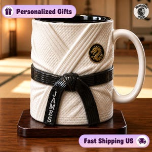 Puede incluir: Una taza de cerámica blanca texturizada para parecer un gi de artes marciales, con un cinturón negro envuelto alrededor del centro. El nombre JAMES está impreso verticalmente en el cinturón. Un emblema dorado circular con un dragón está arriba.