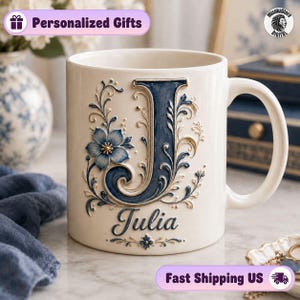 Può includere: Una tazza in ceramica color crema con una grande lettera J in rilievo di colore blu scuro, decorata con intricati motivi floreali e accenti dorati. Sotto la lettera, il nome Julia è scritto in un carattere corsivo blu scuro con dettagli.