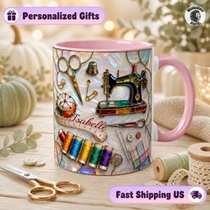 May include: A ceramic mug with a pink handle and interior, featuring a stained-glass style print of sewing tools including a sewing machine, scissors, thimble, measuring tape, and spools of thread, with the name Isabelle printed in script.