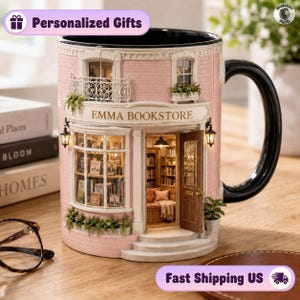 Puede incluir: Una taza de cerámica con forma de librería de ladrillo rosa con el texto EMMA BOOKSTORE sobre la entrada. La taza cuenta con un asa negra, una fachada detallada con escaparate, un balcón y una puerta abierta que revela el interior.