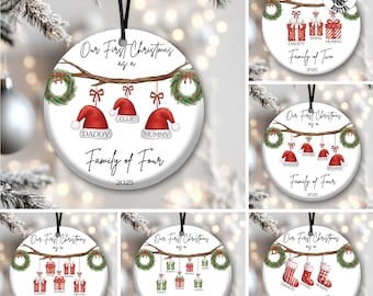 May include: White ceramic Christmas ornament with a black ribbon. The ornament features the text "Our First Christmas as a Family of Four 2025". It includes Santa hats and wreaths. The ornament is hanging from a Christmas tree.