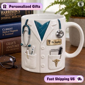 Puede incluir: Una taza de cerámica blanca con forma de bata de médico y una camisa azul verdoso debajo. Presenta un estetoscopio esculpido, una placa con el nombre Dr. Sophia, un bloc de notas, bolígrafos, un termómetro y un emblema de caduceo dorado.