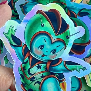 May include: A holographic sticker featuring a cartoon astronaut with a green and blue spacesuit, a red belt, and a star on the chest. The sticker has the text "© 2014 STACEY MARTIN TATTOOS" on the bottom right corner.
