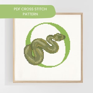 May include: A framed cross-stitch pattern showing a green snake inside a green circle. The snake is detailed with shades of green and brown. The text "PDF CROSS STITCH PATTERN" is in a green banner at the top.