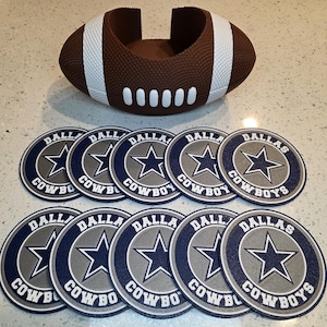 May include: Set of ten round coasters featuring the Dallas Cowboys logo. The coasters are blue and gray with a white star and the words "Dallas Cowboys". The set also includes a brown football-shaped coaster holder.
