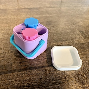 May include: A square lavender container with a bright blue handle, featuring two small circular inserts with scalloped blue and pink lids inside. A separate white square lid sits to the right of the main container on the surface.