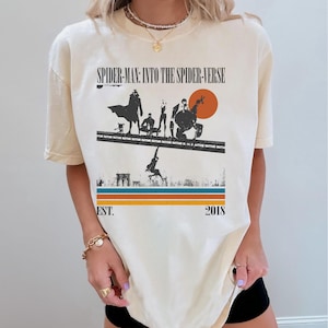 Puede incluir: Camiseta color crema con un gr&aacute;fico de "Spider-Man: Into the Spider-Verse". El dise&ntilde;o incluye siluetas de personajes, un paisaje urbano y el texto "EST. 2018". La camiseta tiene un esquema de color retro con rayas naranjas, azules y marrones.