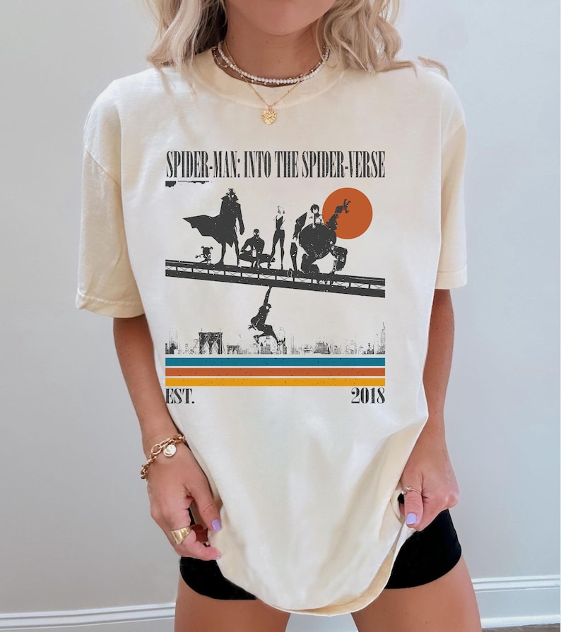 Puede incluir: Camiseta color crema con un gr&aacute;fico de "Spider-Man: Into the Spider-Verse". El dise&ntilde;o incluye siluetas de personajes, un paisaje urbano y el texto "EST. 2018". La camiseta tiene un esquema de color retro con rayas naranjas, azules y marrones.