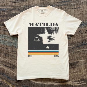 May include: Off-white t-shirt with a graphic of a woman's face and the word "MATILDA" above. Below the image are horizontal stripes in blue, orange, and brown, with "EST. 1996" printed below. The shirt has short sleeves and a crew neck.