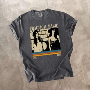 Puede incluir: Camiseta gris oscuro con un gráfico que dice "PRACTICAL MAGIC" encima de una imagen de dos mujeres. Debajo de la imagen están las palabras "EST. 1998" y tres rayas horizontales en azul, naranja y marrón.