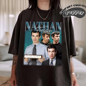 Pode incluir: Camiseta preta com um gráfico de quatro homens. O gráfico inclui o texto "Nathan Fielder" e uma placa de identificação que diz "Nathan Fielder, Winner Ever".